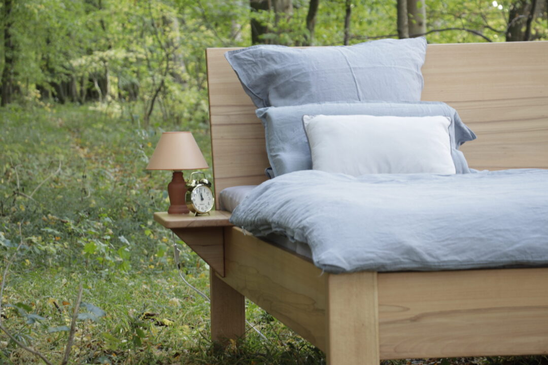 Ein Biobett mit einer Ablage steht im Wald. Auf der Ablage steht ein Wecker und eine Lampe